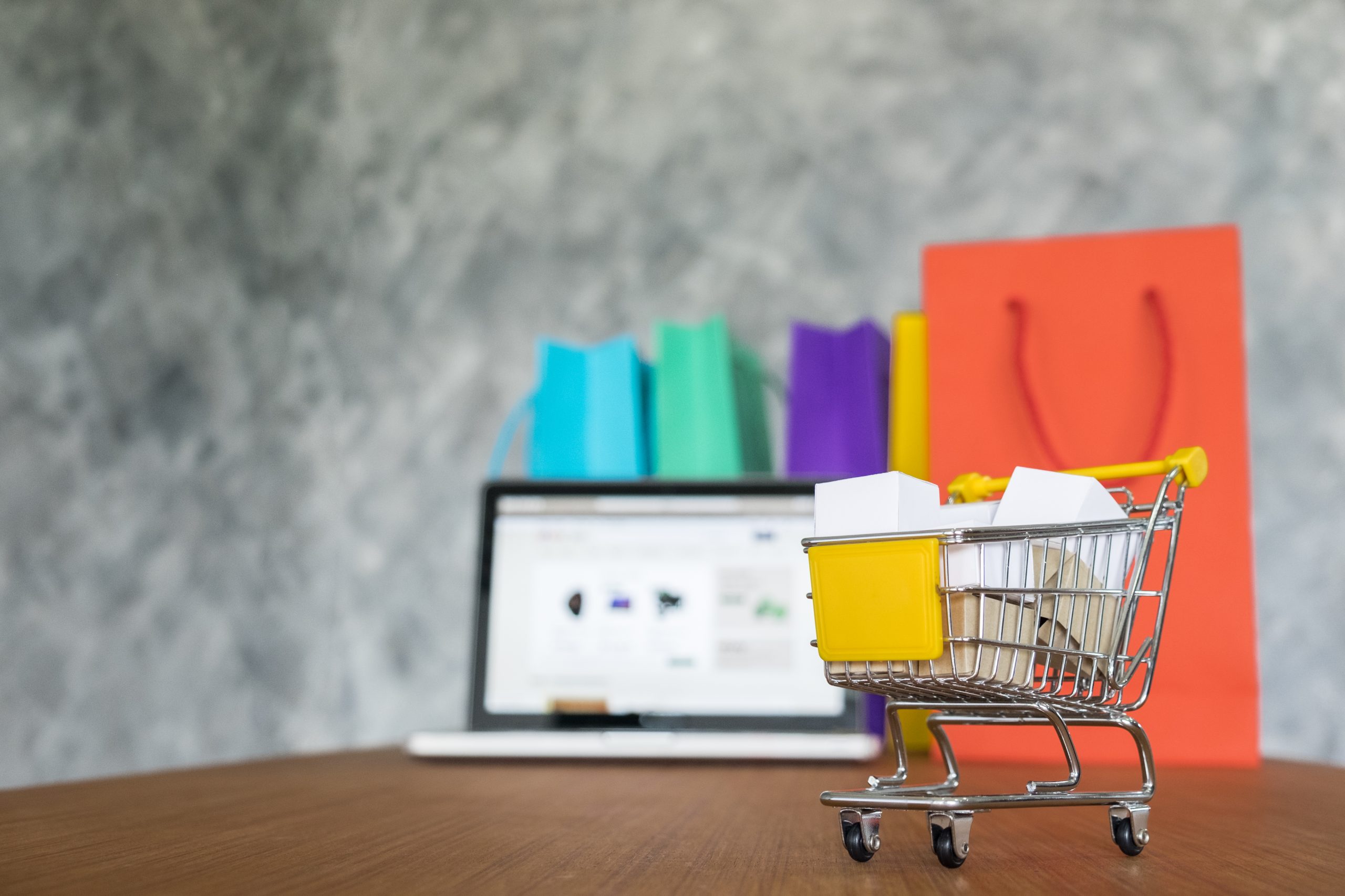 Miniature shopping cart with small boxes displayed in front of a laptop and colorful shopping bags, symbolising online shopping and effective eCommerce SEO strategies.